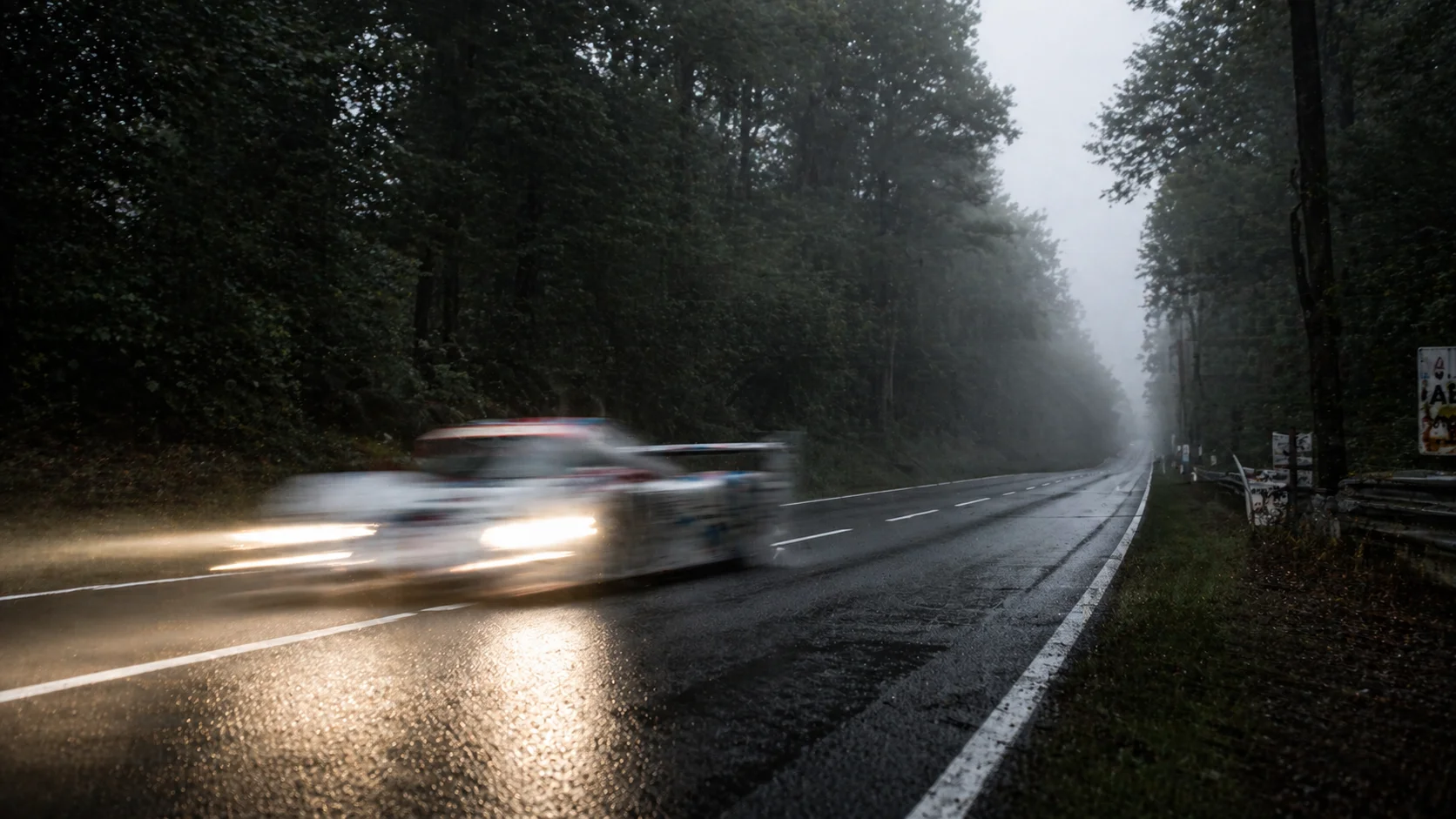 A low-angle blurred action shot of a prototype race car speeding down a narrow forest straight at dawn.