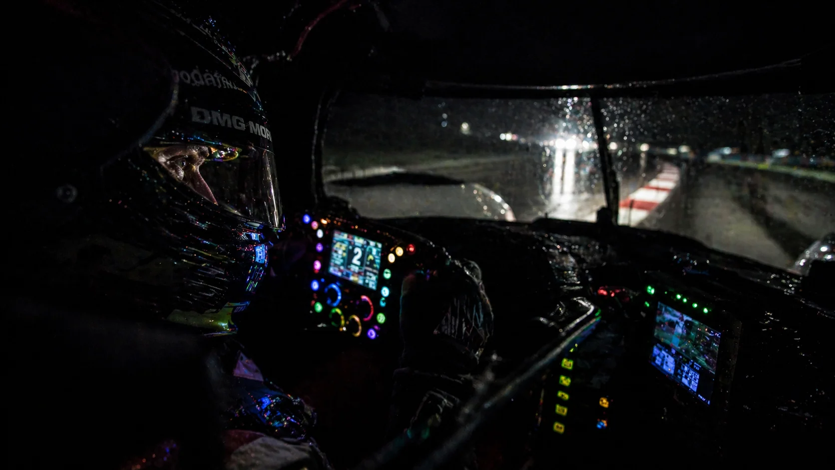 A close-up nighttime shot inside a glowing modern race car cockpit, showing the driver's intense focus in the dark.